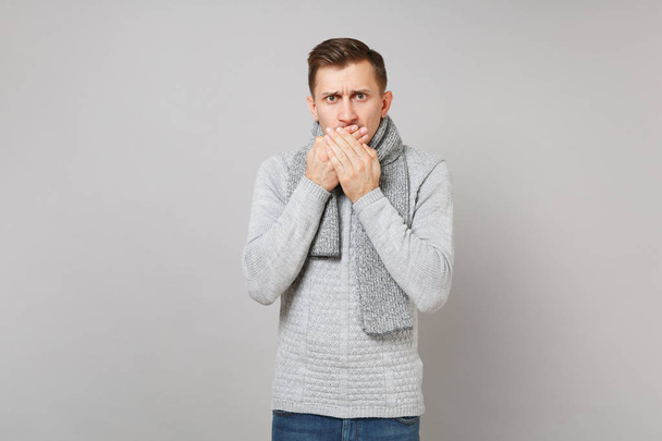 Concerned young man in gray sweater, scarf covering mouth with hands isolated on grey background in studio. Healthy fashion lifestyle, people sincere emotions, cold season concept. Mock up copy space - Foto, Imagen