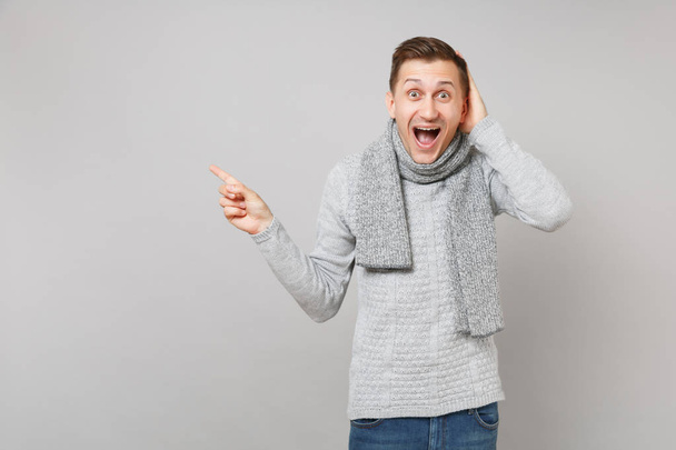 グレーのセーターに驚いた若い男はスカーフ ヘッドの人差し指が脇口を大きく開いて灰色の背景上で分離を維持に手を置く。健康的なライフ スタイルの人々 の感情、寒い季節の概念 - 写真・画像