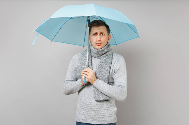Puzzled young man in gray sweater, scarf holding blue umbrella isolated on grey background, studio portrait. Healthy fashion lifestyle people sincere emotions, cold season concept. Mock up copy space - Photo, Image