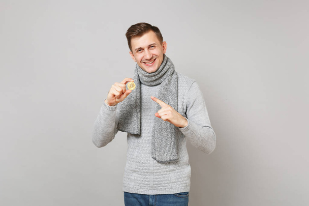 Young man in gray sweater, scarf pointing index finger on bitcoin, future currency isolated on grey wall background. Healthy lifestyle people sincere emotions, cold season concept. Mock up copy space - Foto, imagen