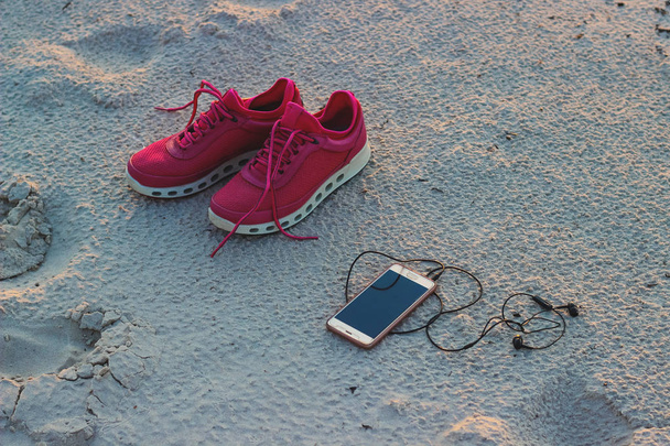 Sports set. Pink sneakers and a mobile phone with headphones close-up on a sandy beach by the sea during sunset. - Foto, Imagem