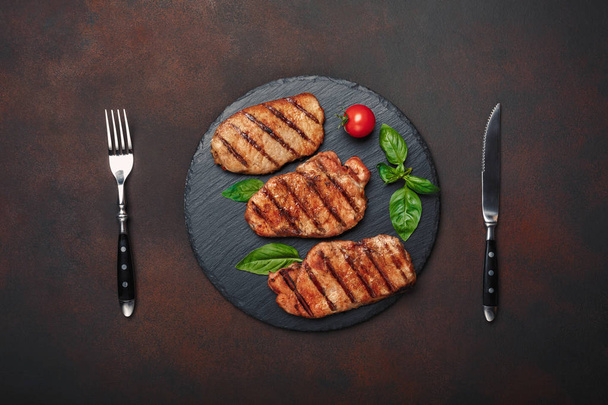 Steaks de porc grillés au basilic, tomates, couteau et fourchette sur pierre noire et fond brun rouillé. Vue du dessus
. - Photo, image