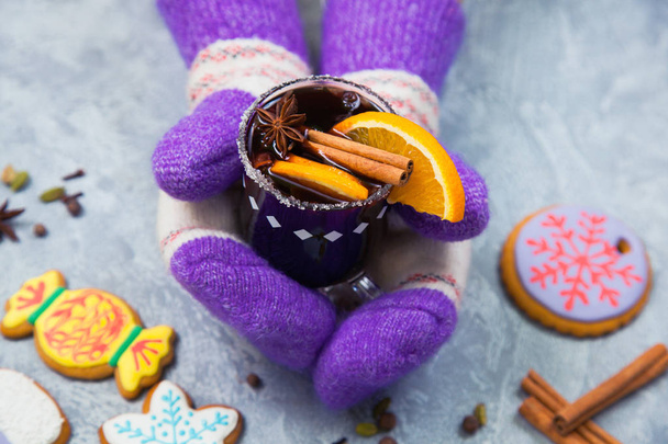 Mulled wine in glass mug with spices, female hands in knitted mittens. Christmas hot drink with decorated new year ginger bread on gray stone table. Top view. - Zdjęcie, obraz