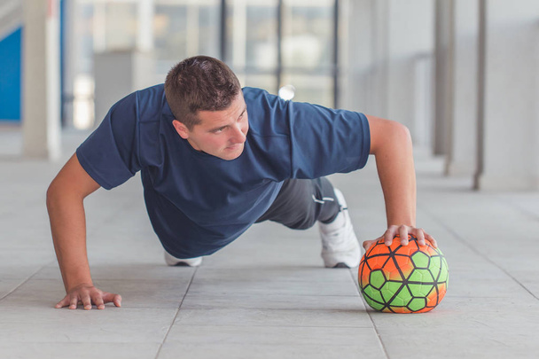 Joven atlético haciendo flexiones con pelota de fútbol en un centro deportivo
  - Foto, Imagen