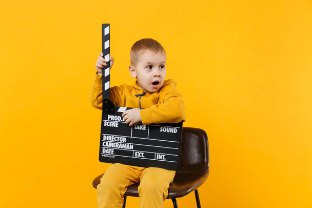 Little kid boy 3-4 years old in yellow clothes isolated on orange wall background, children studio portrait. People childhood lifestyle concept. Hand hold film making clapperboard. Mock up copy space - Photo, image
