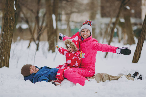 Сміється сімейна жінка, чоловік маленька дівчинка в теплому одязі лежить, сидить на снігу, розсовуючи руки в парку або лісі на відкритому повітрі. Зимові розваги, дозвілля у відпустці. Любов стосунки люди концепція способу життя
 - Фото, зображення