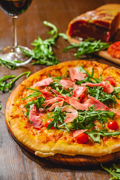 Italian pizza with prosciutto (parma ham), arugula (salad rocket) and cherry tomatoes on wooden board. Close up - Foto, afbeelding