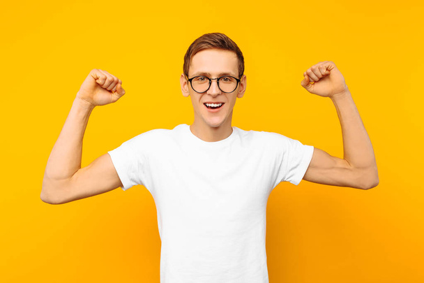 Happy man in glasses, dressed in a white T-shirt, the man who won shows a gesture of victory and success, on a yellow background - Фото, изображение
