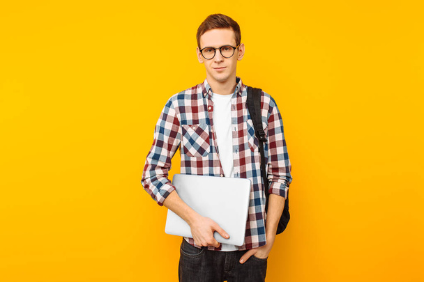 Glücklicher Typ mit Brille, Student im karierten Hemd, mit geschlossenem Laptop in der Hand, auf gelbem Hintergrund - Foto, Bild