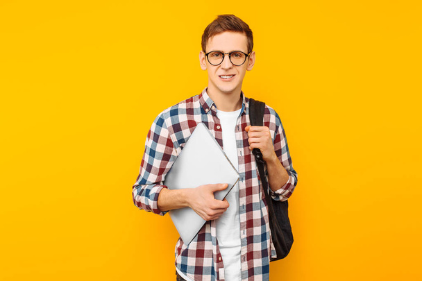 Happy guy with glasses, a student in a plaid shirt, with a closed laptop in hand, on a yellow background - Fotó, kép