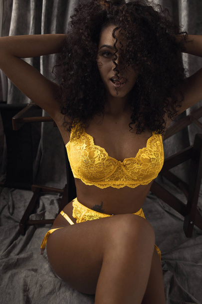 curly-haired brunette sits on the floor in yellow underwear lace and sexually looking at the camera sticking out the tongue - Foto, Imagem