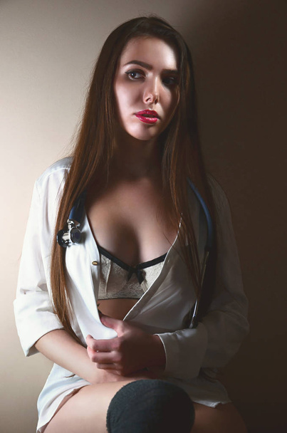 Enfermeira jovem e atraente em uma camisa branca e roupa interior branca com um estetoscópio em torno de seu pescoço sentado em uma sala com paredes bege. Estudante caucasiano sexy na escola de medicina
 - Foto, Imagem