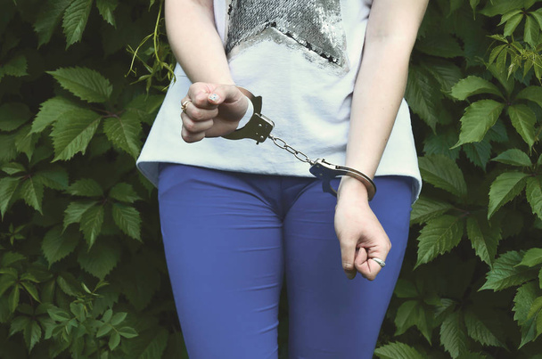 Fragmento do corpo de uma jovem criminosa com as mãos algemadas contra um fundo de folhas de hera florescente verde. O conceito de detenção de uma criminosa num ambiente rural
 - Foto, Imagem