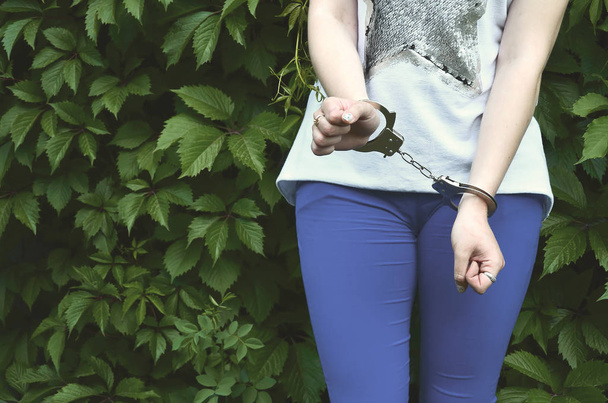 Фрагмент тіла молодої кримінальної дівчини з руками в наручниках на фоні зеленого квітучого листя плюща. Концепція затримання злочинця жінки-злочинця в сільському середовищі
 - Фото, зображення