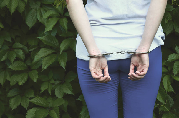 Fragment of a young criminal girl's body with hands in handcuffs against a green blossoming ivy leaves background. The concept of detaining an offender of a female criminal in a rural environment - Photo, Image