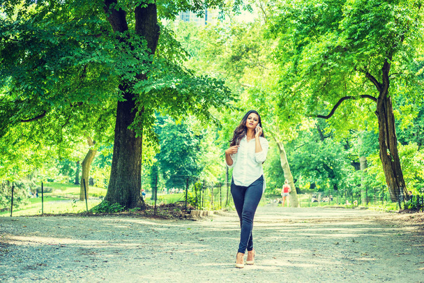Fiatal indiai-amerikai nő utazik, a Central Park, New York, pihentető visel, fehér ing, fekete nadrág, magas sarkú, séta a road, a zöld fák, forró nyár, mobiltelefonon beszél - Fotó, kép