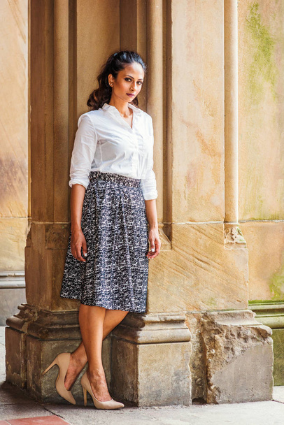 Portrait of Young Beautiful East Indian American Woman in New York, wearing white shirt, black and white patterned skirt, high heals, standing by vintage column, turning around, looking at you - Photo, image