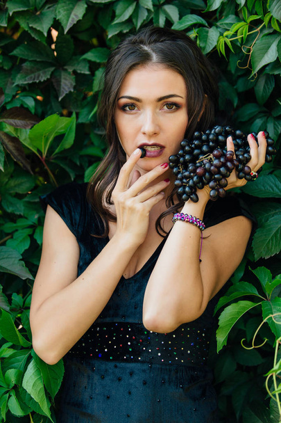 Portrait of young sexy brunette in black dress biting black grapes. - Foto, afbeelding