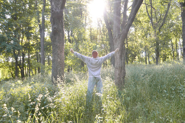 Gri spor elbiseli bir genç adam ormandaki ağaçlar arasında güneşin düşüyor sevinir. Rekreasyon spor sırasında açık hava ormanda çalıştırın. Dawn güzel zevk - Fotoğraf, Görsel