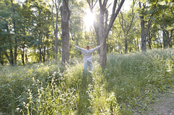 Ένας νεαρός άντρας σε ένα κοστούμι γκρι αθλητικά χαίρεται με την άνοδο του ήλιου ανάμεσα στα δέντρα στο δάσος. Ψυχαγωγία κατά τη διάρκεια αθλητικών τρέχει στο δάσος ανοιχτό αέρα. Η χαρά του μια όμορφη αυγή - Φωτογραφία, εικόνα