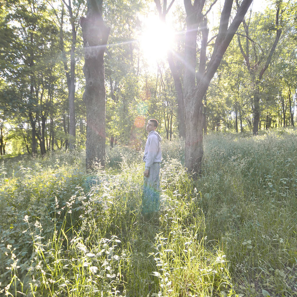 Молодой парень в сером спортивном костюме стоит напротив солнца среди деревьев в лесу. Отдых во время спортивных пробежек в лесу под открытым небом. Восхищение прекрасным рассветом
 - Фото, изображение
