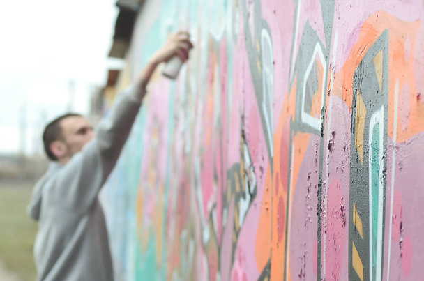 A young guy in a gray hoodie paints graffiti in pink and green colors on a wall in rainy weather. Focus on the fragment of wall and blurred artist - Photo, Image