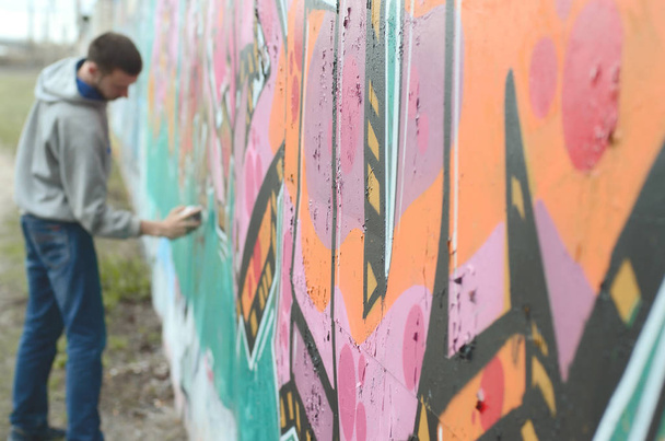 A young guy in a gray hoodie paints graffiti in pink and green colors on a wall in rainy weather. Focus on the fragment of wall and blurred artist - Foto, Bild