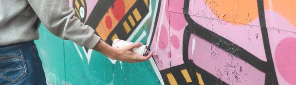 Hand of a young guy in a gray hoodie paints graffiti in pink and green colors on a wall in rainy weather - Zdjęcie, obraz