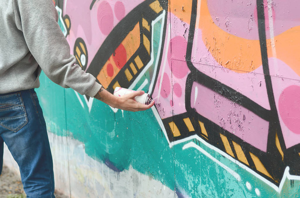Hand of a young guy in a gray hoodie paints graffiti in pink and green colors on a wall in rainy weather - Photo, Image