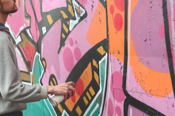 Mladý muž v šedém hoodie maluje graffiti v růžové a zelené barvy na zeď v deštivém počasí - Fotografie, Obrázek