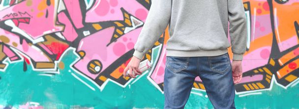 A young graffiti artist in a gray hoodie looks at the wall with his graffiti in pink and green colors on a wall in rainy weather. Street art concept - Foto, Imagen