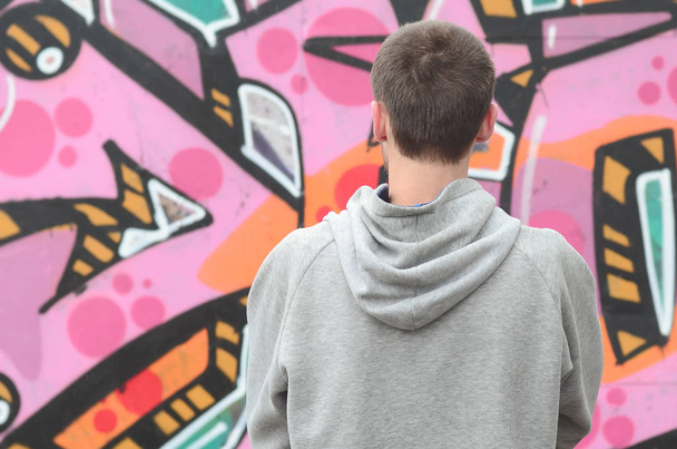 Egy fiatal graffiti művész egy szürke kapucnis úgy néz ki a fal a graffiti, rózsaszín és zöld színben a falra, az esős időjárás. Utcai művészeti koncepció - Fotó, kép