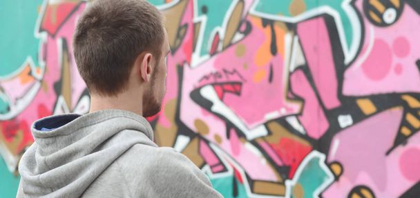 Ένας καλλιτέχνης νέος γκράφιτι σε ένα γκρι φούτερ με κουκούλα εξετάζει τον τοίχο με του γκράφιτι σε ροζ και πράσινο χρώμα σε έναν τοίχο στο βροχερό καιρό. Τέχνη του δρόμου έννοια - Φωτογραφία, εικόνα