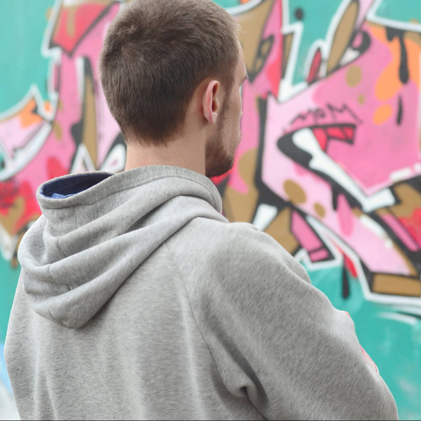 Nuori graffititaiteilija harmaassa hupparissa katsoo seinää graffitilla vaaleanpunaisissa ja vihreissä väreissä sateisessa säässä. Katutaiteen käsite
 - Valokuva, kuva