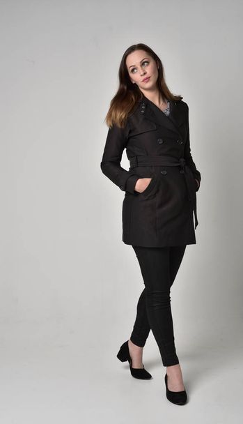 full length portrait of a brunette girl wearing long black coat, standing pose on grey studio background. - Foto, Imagem