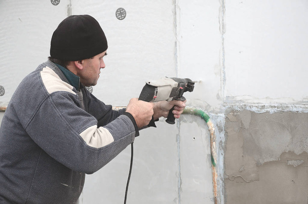 Un operaio anziano fa un foro in una parete di polistirolo per la successiva installazione di un tassello di rinforzo in plastica. Creazione di fori nel muro con un trapano. Riscaldamento della facciata dell'edificio
 - Foto, immagini