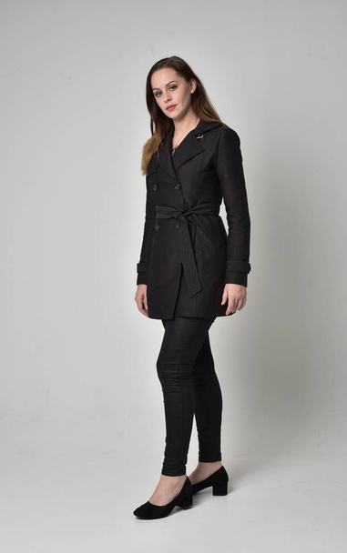 Ολόσωμο πορτρέτο της μια μελαχρινή κοπέλα φορώντας μακρύ μαύρο παλτό, στέκεται πόζα σε φόντο γκρι στούντιο. - Φωτογραφία, εικόνα