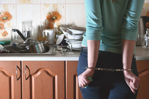 Дівчина в наручниках, незадоволена і втомилася від щоденного миття посуду, стоїть поруч з кухонною стійкою і раковиною, яка заповнена стійкою немитого посуду. Кухня рабство
 - Фото, зображення