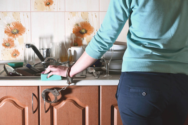 Dívka s houbou v ruce, pouta na kuchyňské lince s umyvadlem s partou neumyté nádobí. Ženské otroctví kuchyně. Problém každodenní ruční mytí nádobí - Fotografie, Obrázek