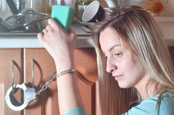 Portrét dívky smutný a unavený, s houbou, spoutaný na kuchyňské lince se spoustou neumyté nádobí, talíře, šálky a příbory. Koncept nutí ženy pro práci v kuchyni - Fotografie, Obrázek