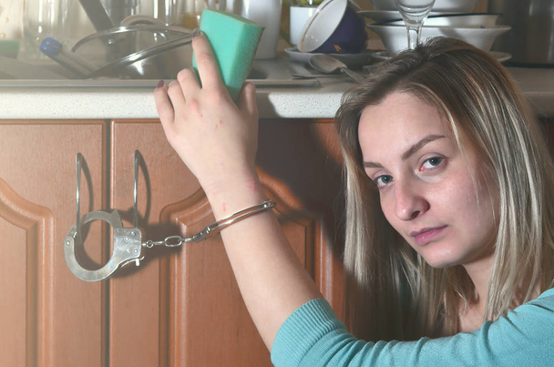 Portrait d'une fille triste et fatiguée avec une éponge, menottée à un comptoir de cuisine avec beaucoup de vaisselle, assiettes, tasses et couverts non lavés. Le concept de forcer les femmes à travailler dans les cuisines
 - Photo, image