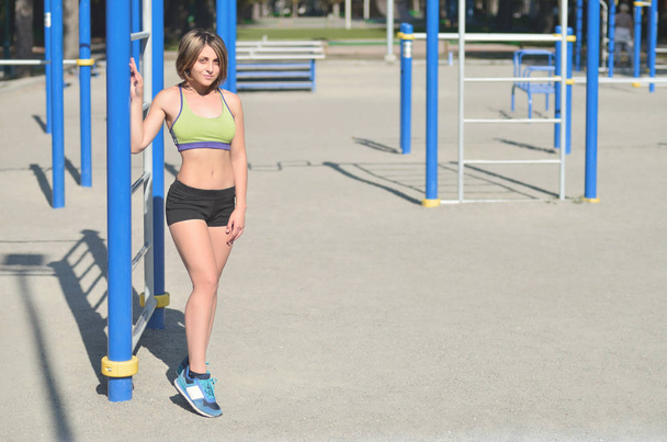 Молода біла дівчина-спортсменка в яскраво-зеленому спортивному одязі стоїть на тлі вуличного спортивного майданчика для тренувань на відкритому повітрі. Літні види спорту та концепція здорового способу життя
 - Фото, зображення