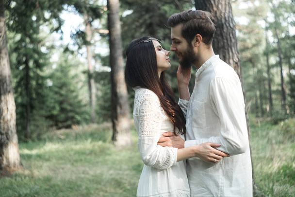 Young beautiful Slavic couple in love walks through the woods, sweet girl in a simple boho-style wedding dress. They are happy and calm. Golden hour, summer day, warm. They kiss, hug and hold hands. - Valokuva, kuva