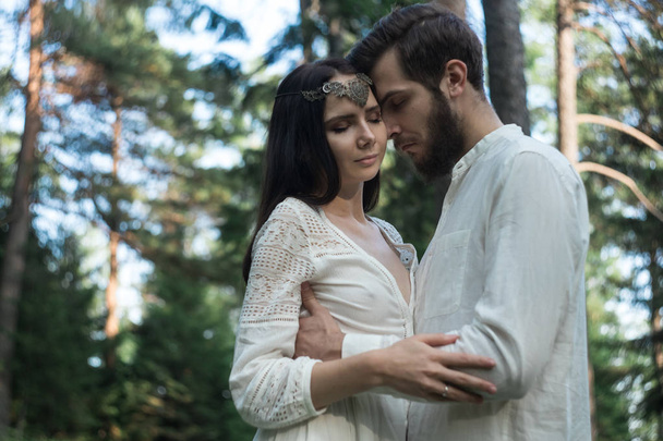 Young beautiful Slavic couple in love walks through the woods, sweet girl in a simple boho-style wedding dress. They are happy and calm. Golden hour, summer day, warm. They kiss, hug and hold hands. - Фото, изображение