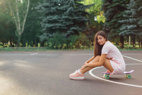 Portrait of a smiling charming brunette female sitting on her skateboard on a basketball court. Happy woman with trendy look taking break during sunset. - Foto, Imagen