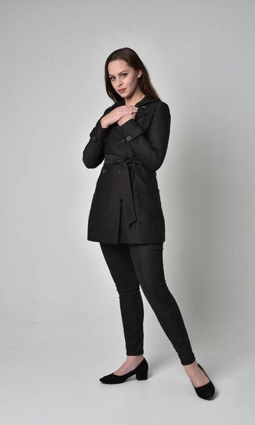 Pełna długość Portret brunetka Girl ubrany długi czarny płaszcz, pozie stojącej na studio szary tło. - Zdjęcie, obraz