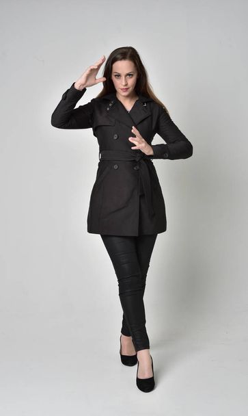 po celé délce portrét bruneta dívka, která nosí dlouhý černý kabát, stojící pozice na šedé studio pozadí. - Fotografie, Obrázek