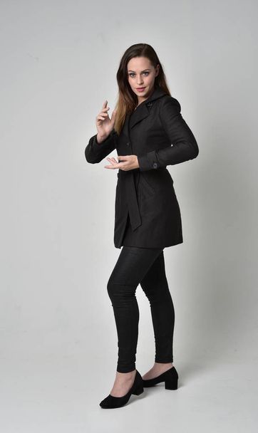full length portrait of a brunette girl wearing long black coat, standing pose on grey studio background. - Фото, изображение