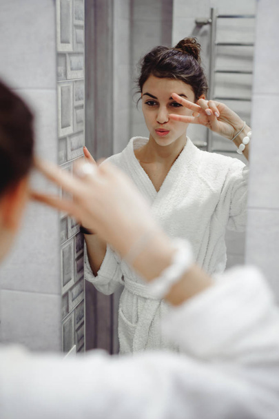 portrait of a beautiful woman in the bathroom. cheerful young girl looks at herself in the mirror, grimaces - Foto, afbeelding