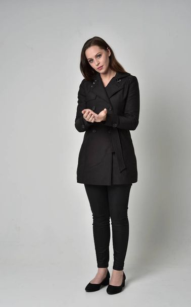 full length portrait of a brunette girl wearing long black coat, standing pose on grey studio background. - Фото, изображение
